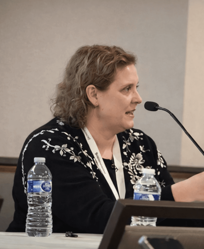 A smiling man in a dark shirt and jeans sits casually on a stool against a dark grey background, looking off to the side.