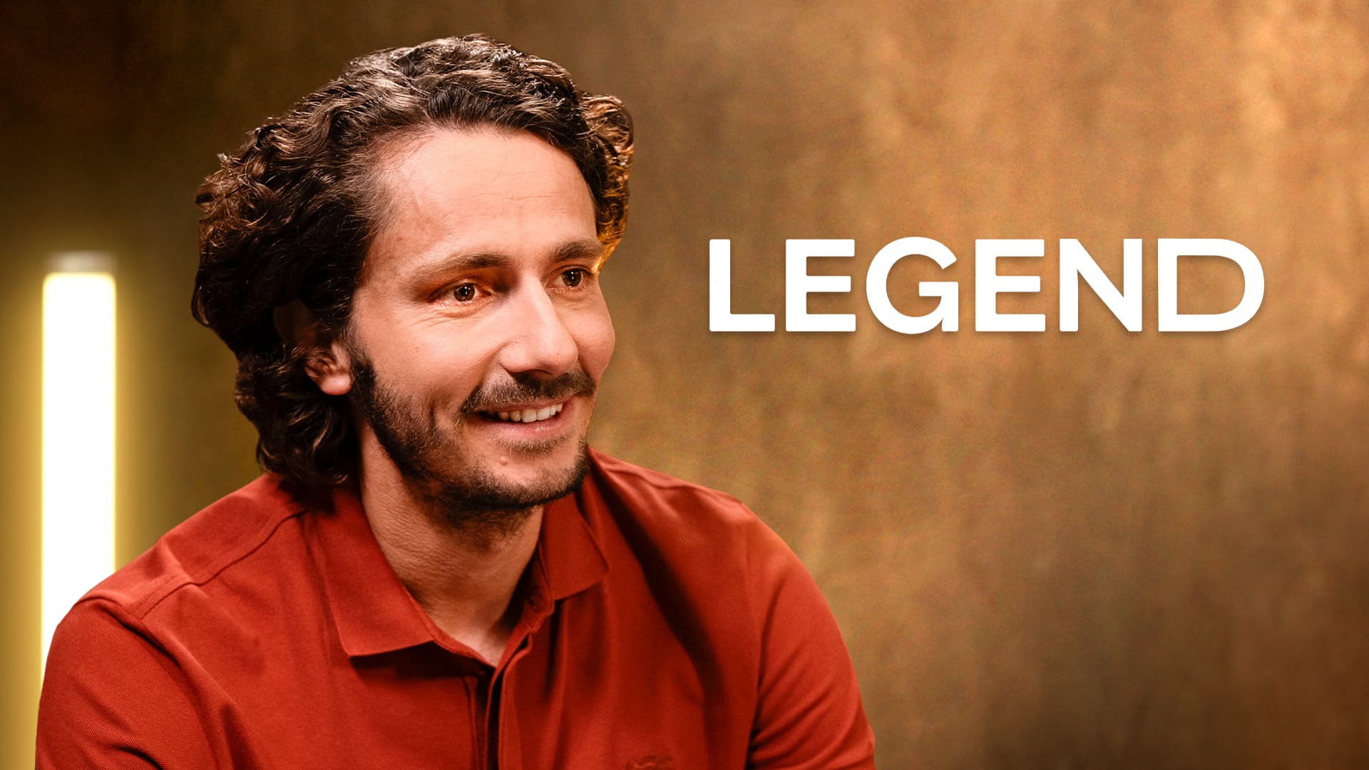 Portrait souriant et professionnel de Guillaume Pley, journaliste et animateur français, fondateur du média digital 'Legend'. Il porte un polo de couleur terre cuite (ou orange brûlé) et est filmé de profil, regardant vers la droite. L'arrière-plan est chaud, flou (bokeh), dominé par des tons or et brun avec une source de lumière verticale en arrière-plan, suggérant un cadre de studio pour une interview ou un podcast. L'image est idéale pour illustrer la collaboration de Nux-Studio avec des figures du digital, notamment dans la stratégie de présence digitale, la production vidéo et la direction artistique de médias en ligne.