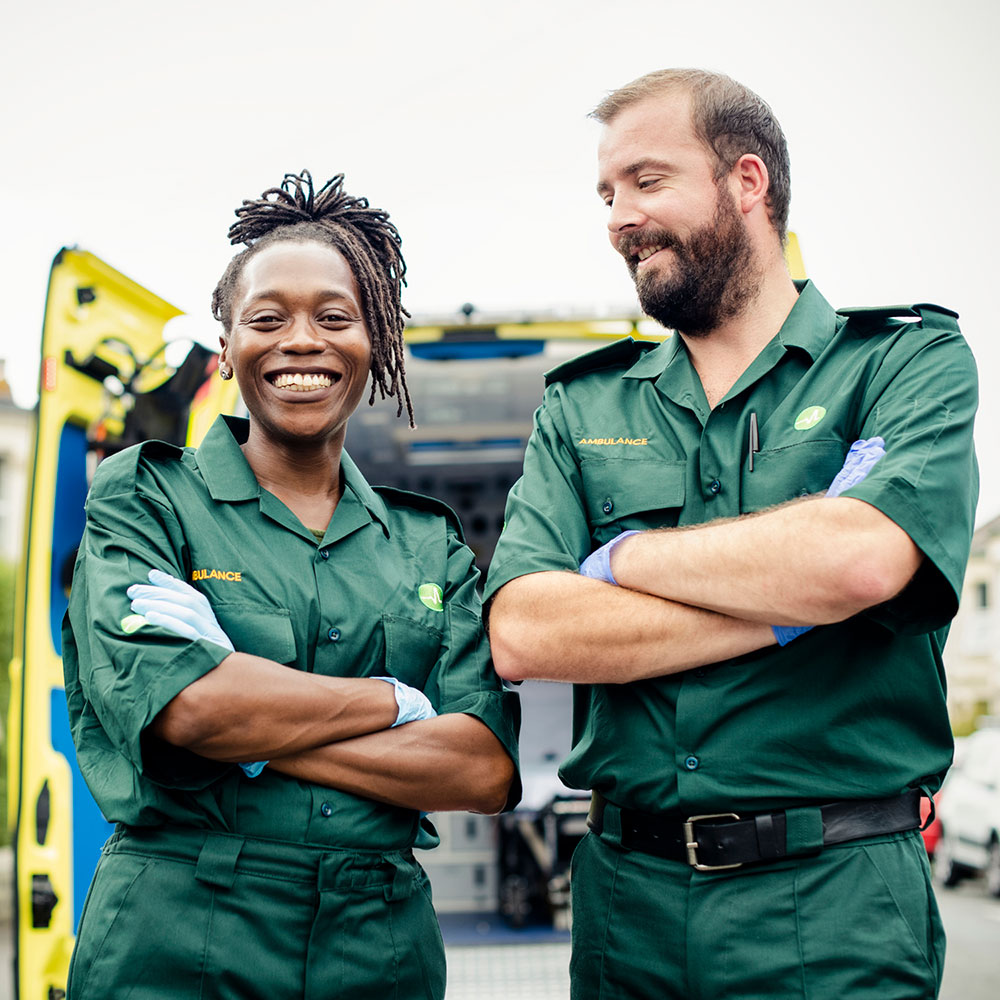 Two paramedics wearing green ambulance service uniforms stand confidently with arms crossed in front of their emergency vehicle. A smiling woman and a bearded man pose together, with medical equipment and their ambulance visible in the background, depicting professional camaraderie and readiness in emergency medical services.