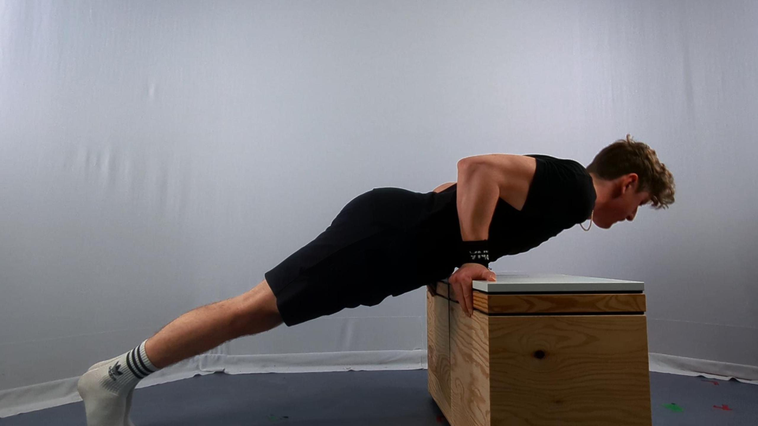 A man in athletic clothing performs calisthenics supinated incline push-ups using a wooden bench against a plain backdrop.