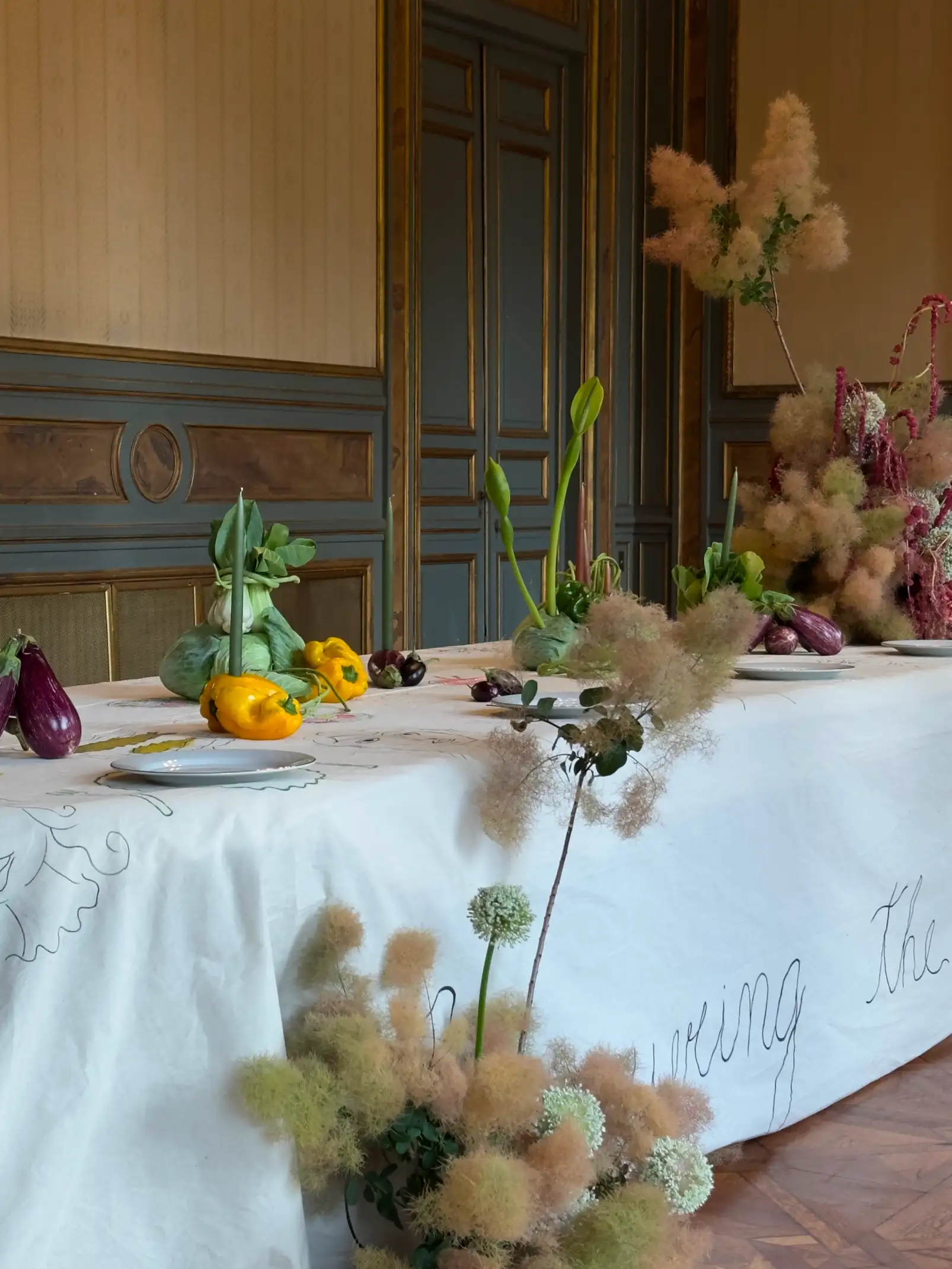 Mesa alargada decorada con un mantel blanco bordado con frases, adornada con berenjenas, repollos, pimientos amarillos y verdes, junto a arreglos florales de hortensias y ramas esponjosas, en un salón elegante con paredes de madera y detalles dorados.