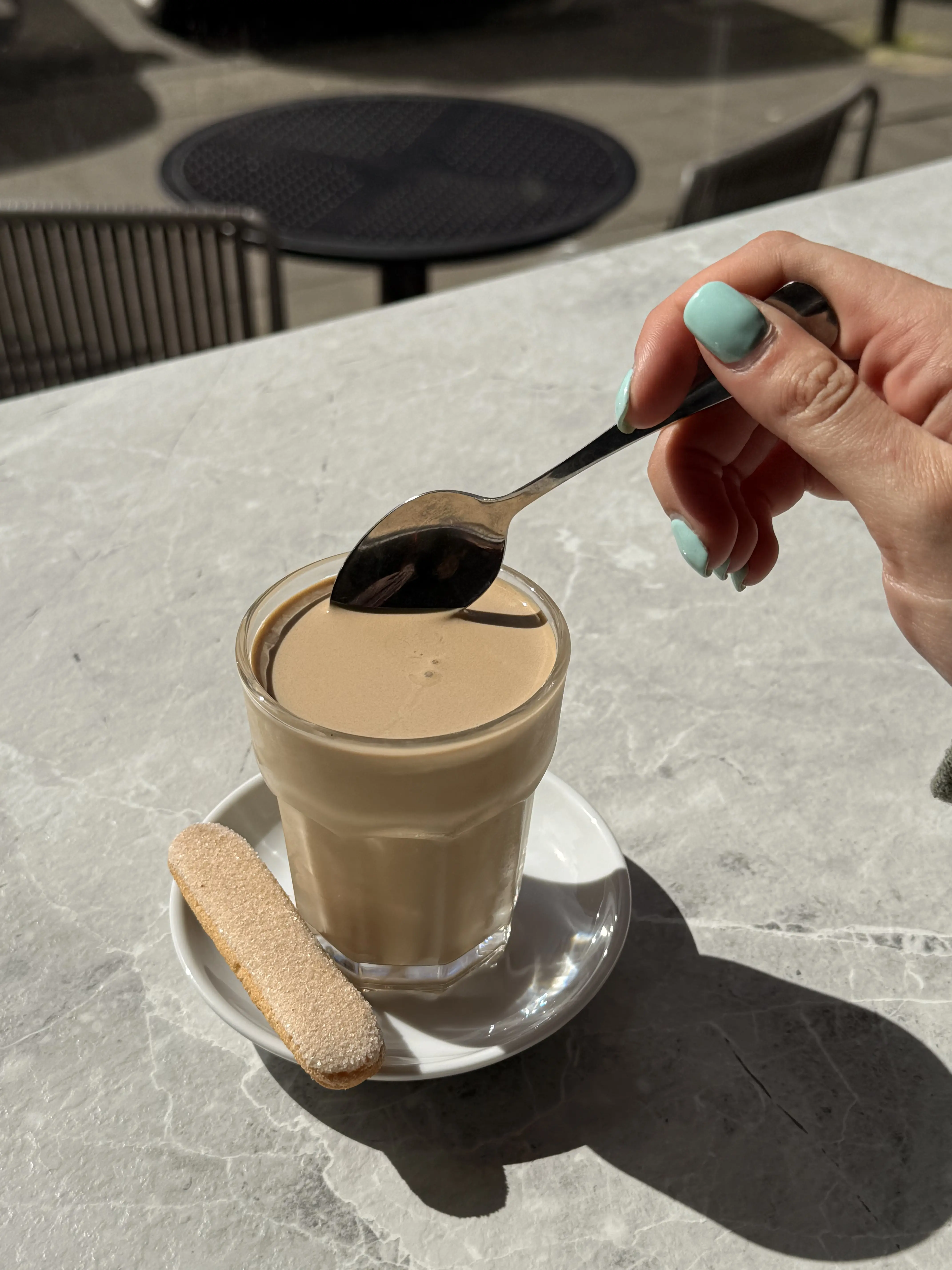 Close-up of a person enjoying a cafe crema with a ladyfinger cookie on a sunny outdoor café table.