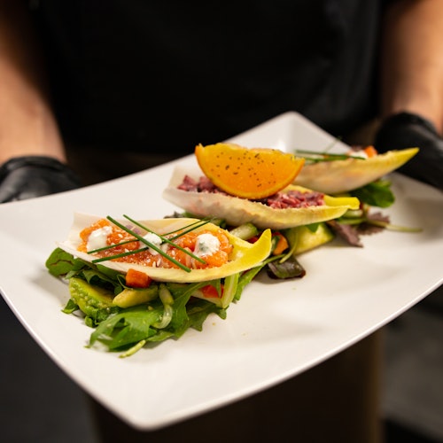 A person is holding a white plate with salad, endive leaves, crumbled cheese, herbs, and an orange wedge garnish.
