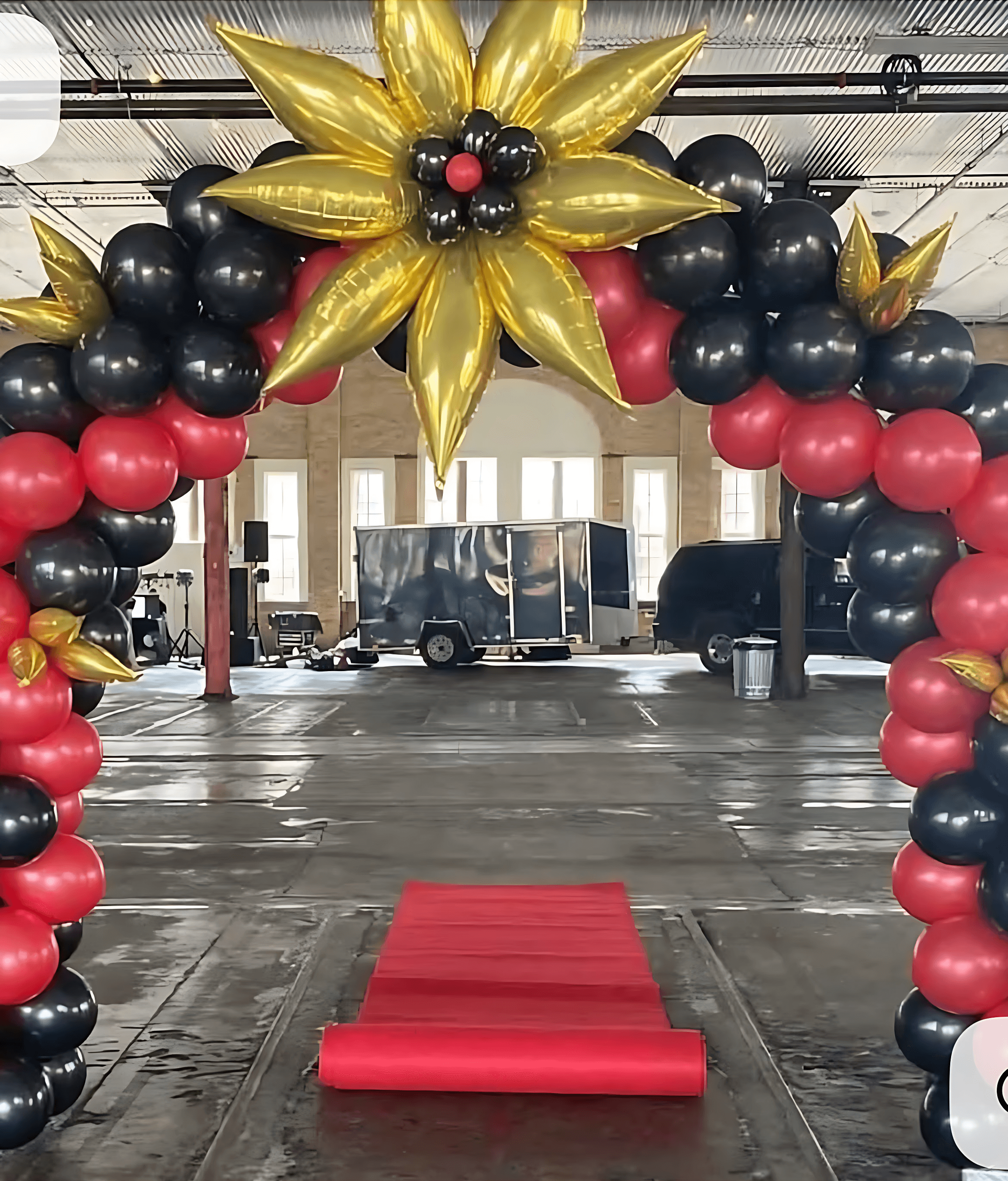 A vibrant balloon arch designed with gold, black, and red balloons in a star pattern towers over a red carpet in an industrial-style venue with vehicles visible in the background.