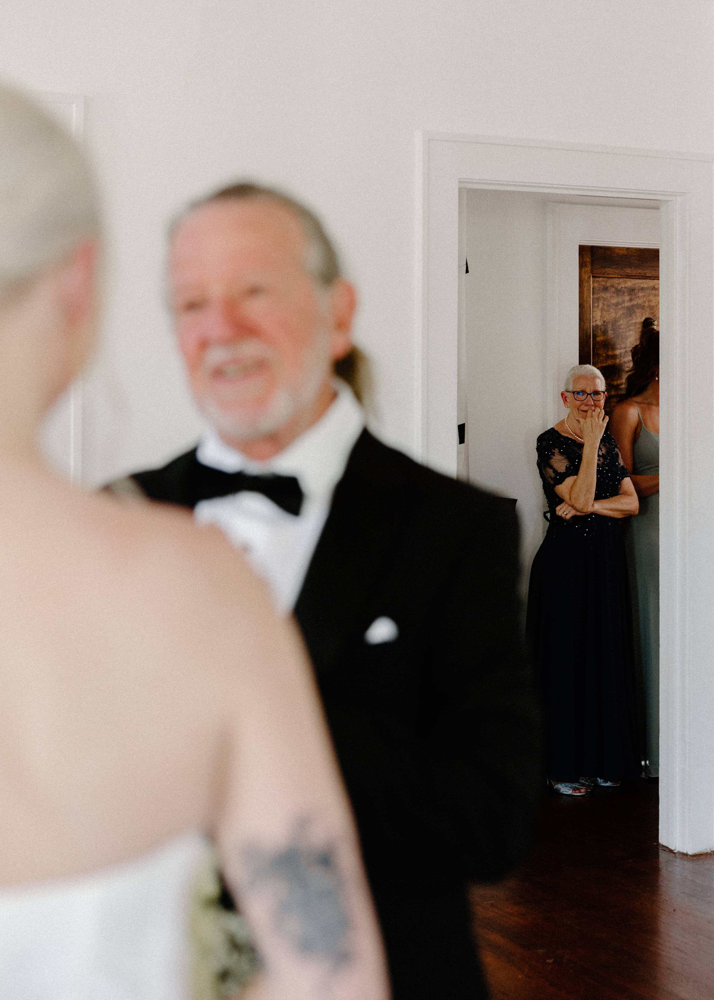 A blurred shot shows a man in a tuxedo smiling at a person in a white dress, possibly a bride. In the background, a person watches from a doorway.