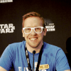 Smiling person in glasses at a panel discussion, wearing a blue shirt, with a backdrop featuring a Star Wars theme.