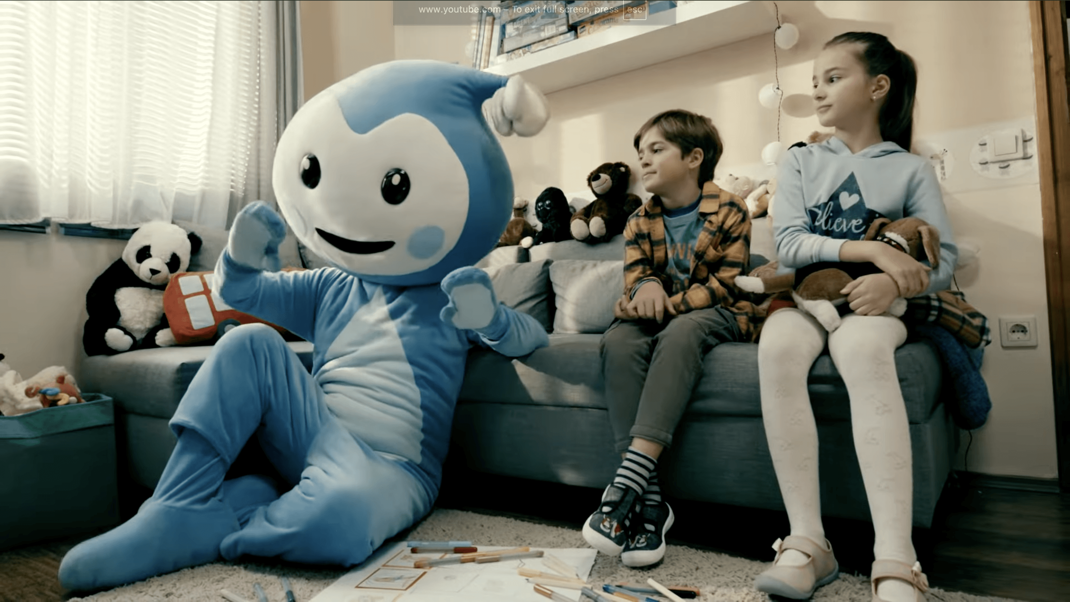 Cseppbarát kékl kabalafigurája a földön ülve énekel a gyerekeknek.