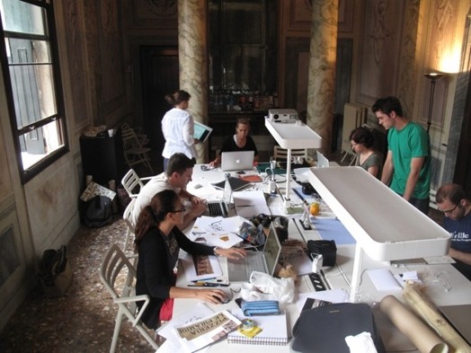 A photo of several students working on laptops at a table while in the middle there is a projector.
