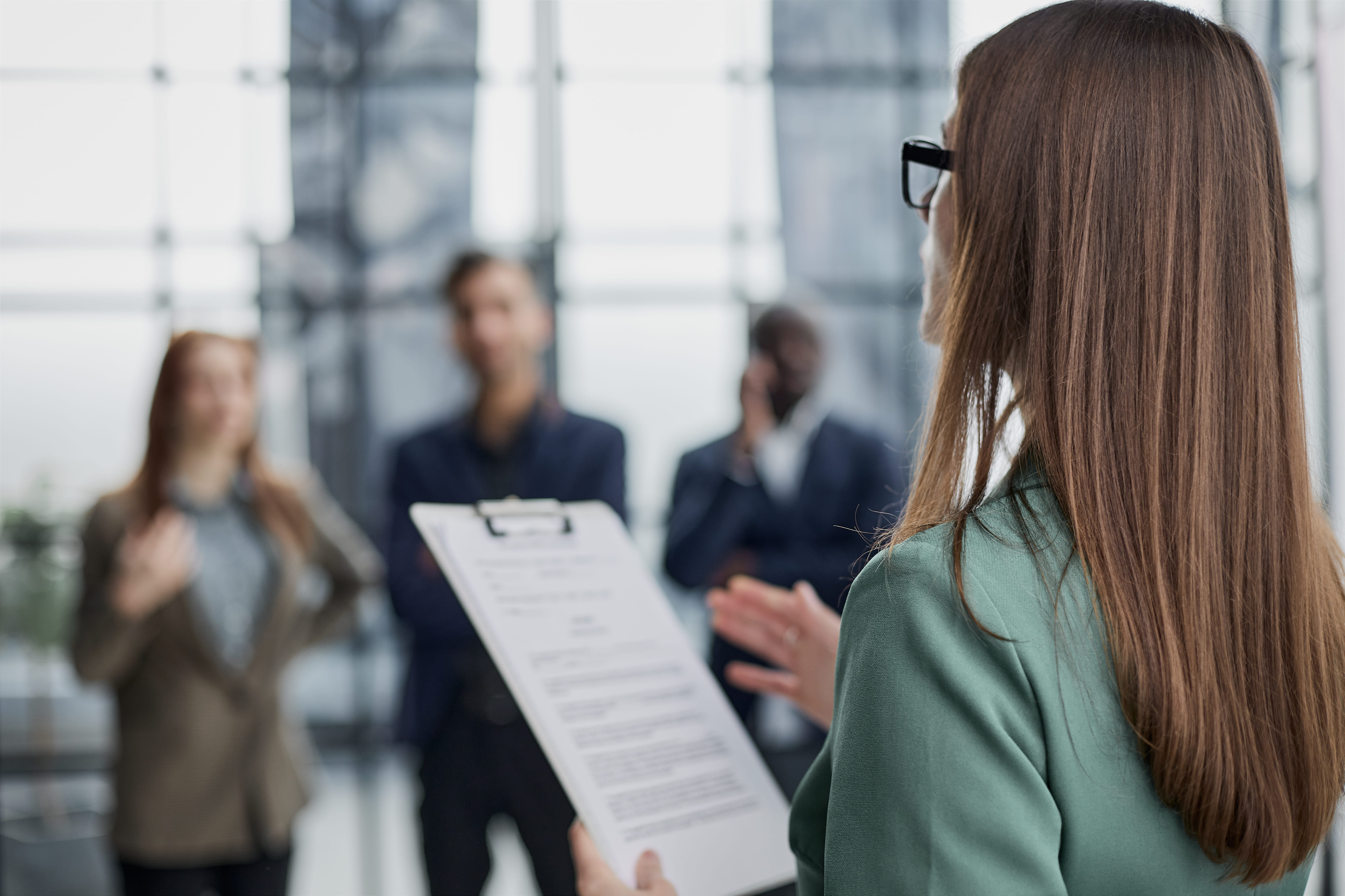 Imagem de uma profissional em um ambiente corporativo segurando uma prancheta com documentos, aparentemente em um momento de apresentação ou briefing para uma equipe. A câmera está posicionada atrás da mulher, destacando seu cabelo longo e sua postura profissional, enquanto os colegas ao fundo, desfocados, estão atentos à sua explicação. O ambiente tem uma aparência moderna, com janelas amplas e boa iluminação natural, sugerindo um ambiente de trabalho colaborativo e profissional.