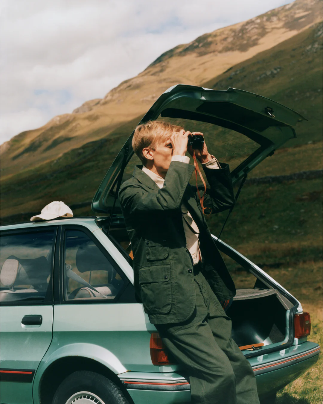 A younge man leaning on a car, looking through binoculars
