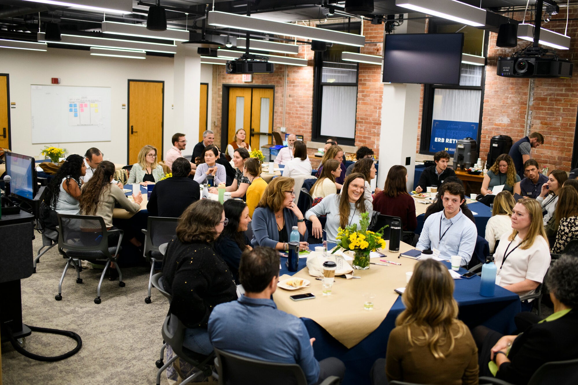 A group of people seated around a large table, engaged in discussion in a casual meeting space.