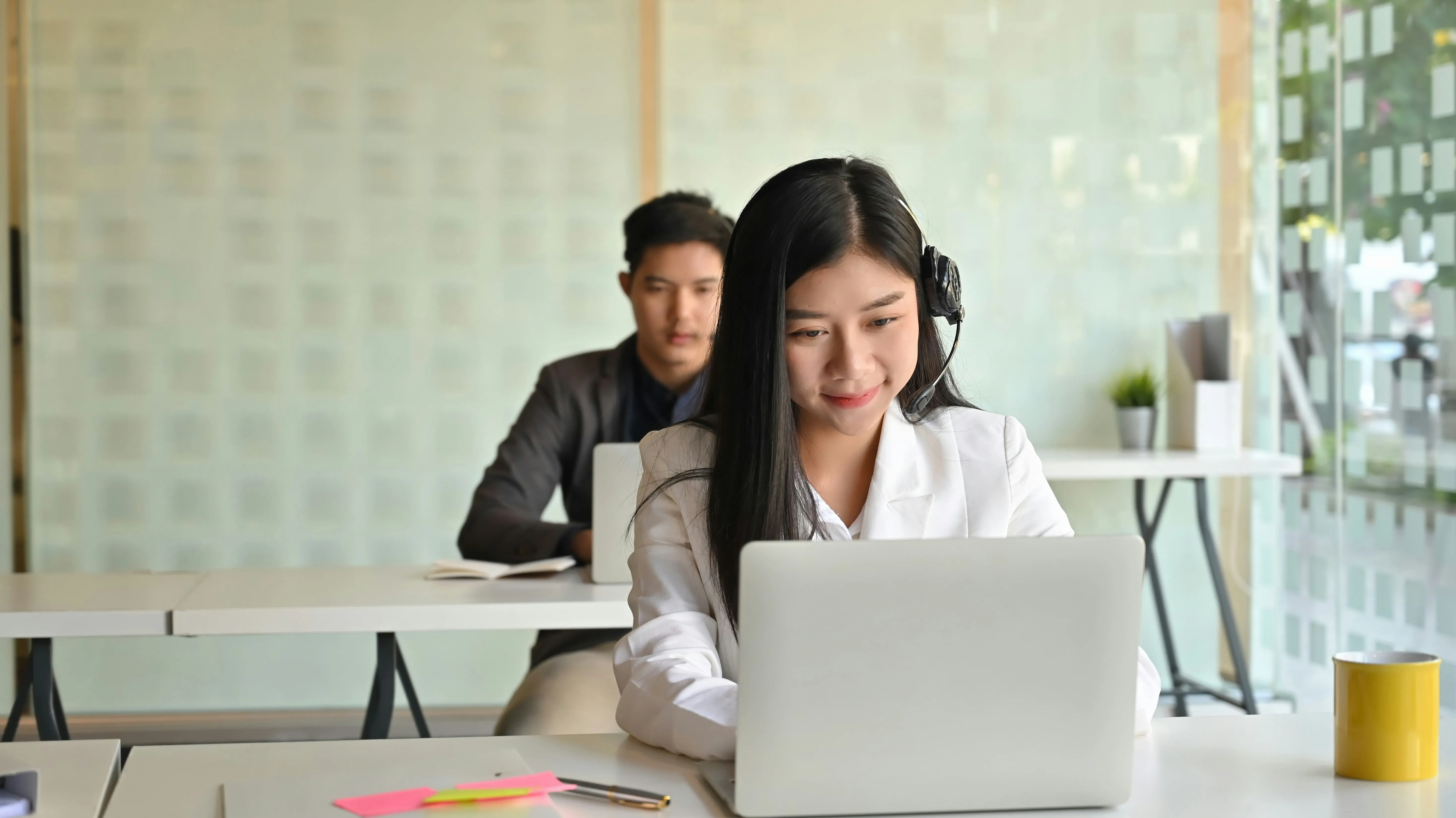 A woman working on a laptop with a focused expression, while a man sits behind her in a bright, modern space.