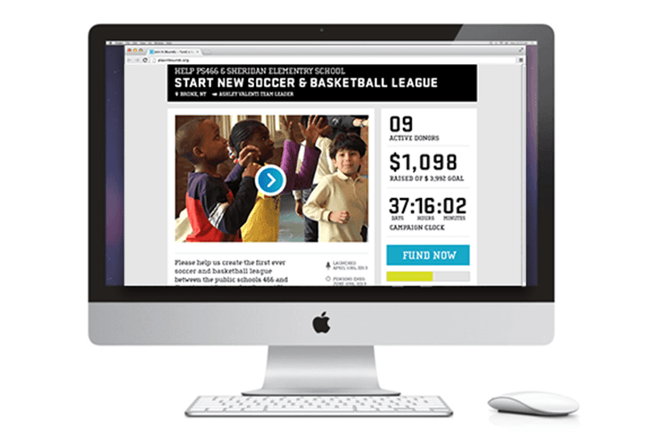 An Apple iMac computer displays a webpage titled “HELP PASS NEW SOCCER & BASKETBALL LEAGUE” from Ashley Valley School. The page shows active donors ($1,098), raised goal ($3,942), and campaign clock (37:16:02). A blue button labeled “FUND NOW” appears below. The webpage includes text encouraging donations to create the first ever soccer and basketball league between public schools 456 and 457. A white Apple keyboard and mouse are positioned in front of the computer.