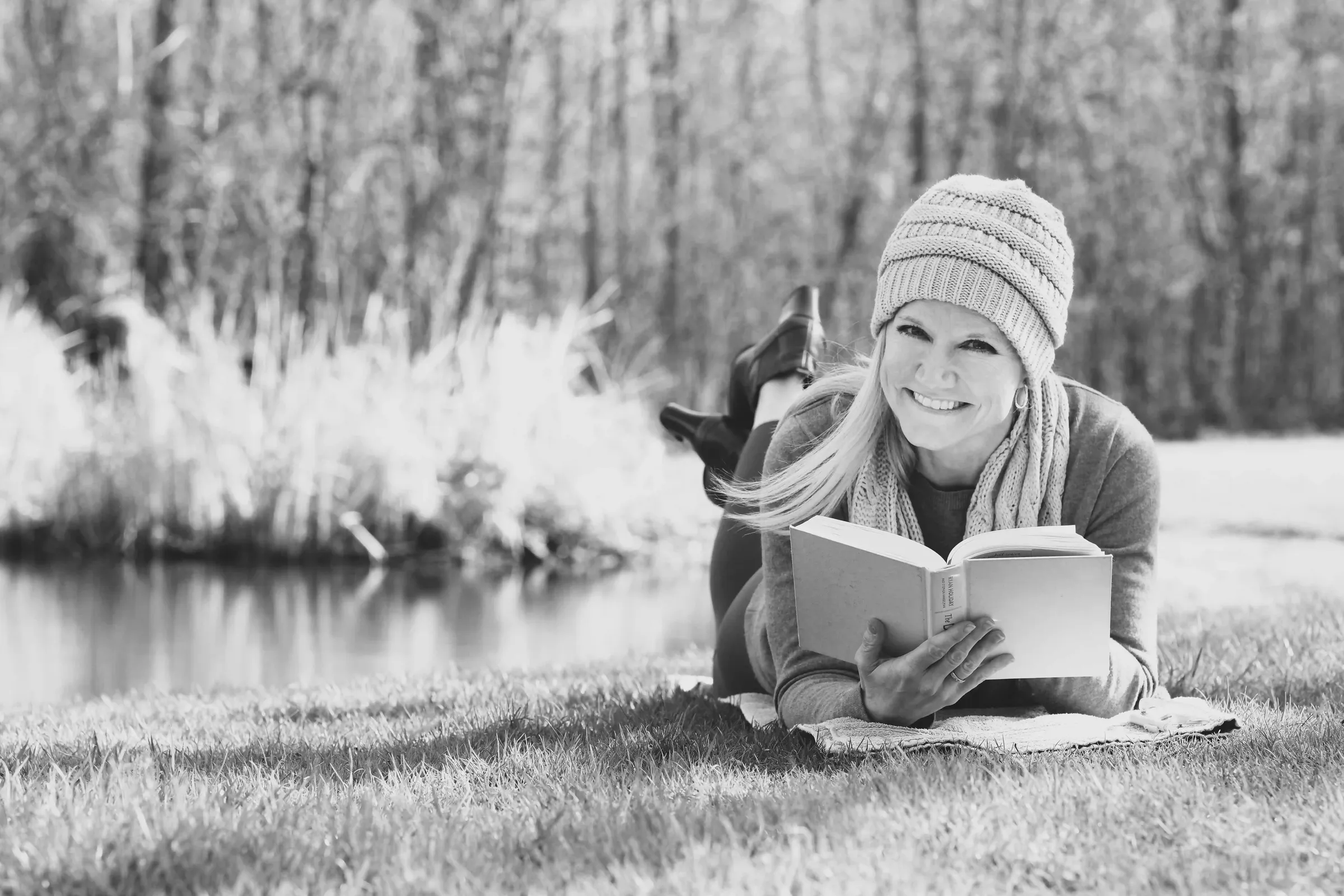 Julie Blank lying on the grass beside a pond, reading a book