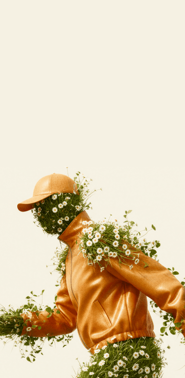 A surrealist digital artwork of a floral figure in a dynamic walking or running pose, shown in profile. The figure wears a textured orange leather jacket and a matching orange cap. Their head, hands, and legs are entirely composed of thick green grass blooming with white daisies, with extra clusters of flowers and green vines sprouting from the shoulders and seams of the jacket against a plain cream background.