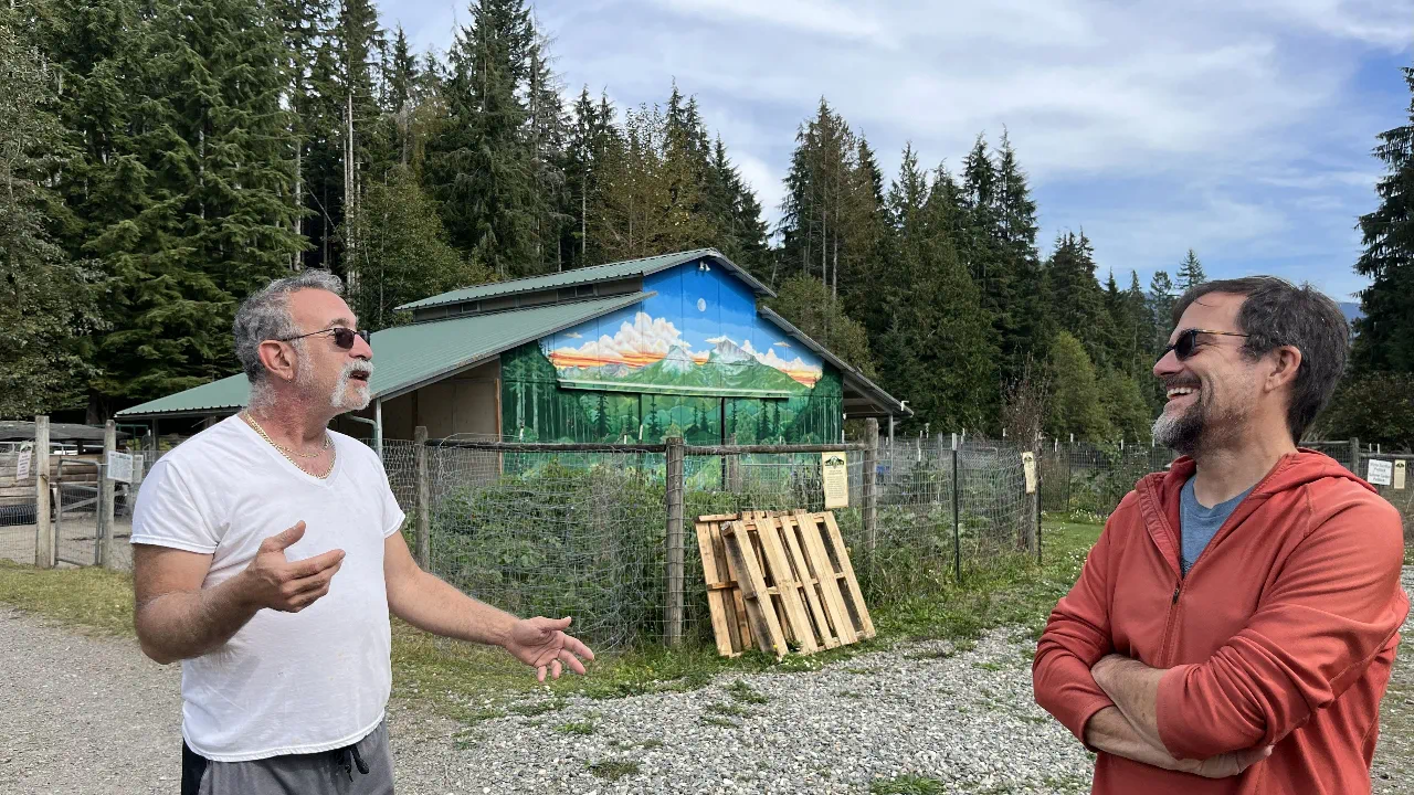 Two community members talking outdoors near a farm building, with forested land and a painted barn in the background.