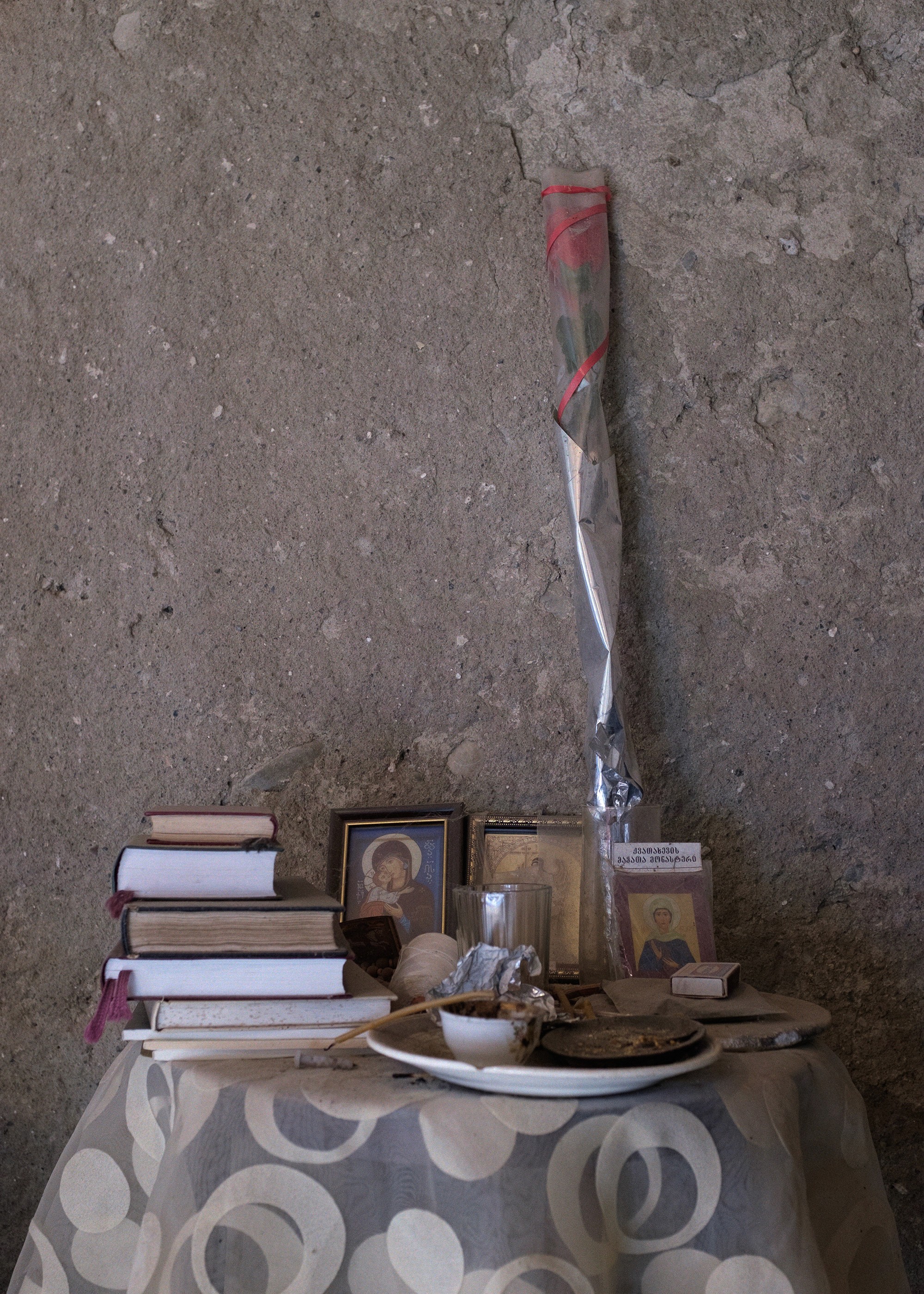 A small memorial table with icons, books, and a single red rose, placed against a concrete wall