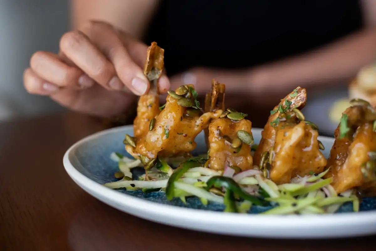A person's hand reaches toward a plate of fried shrimp and sliced cucumbers garnished with green onions.