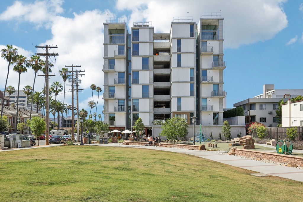 Olive Street Park in Bankers Hill next door to modern, geometric apartment building of Kaya with multiple balconies, set against a bright blue sky. Palm trees, a park, and pathways in the foreground