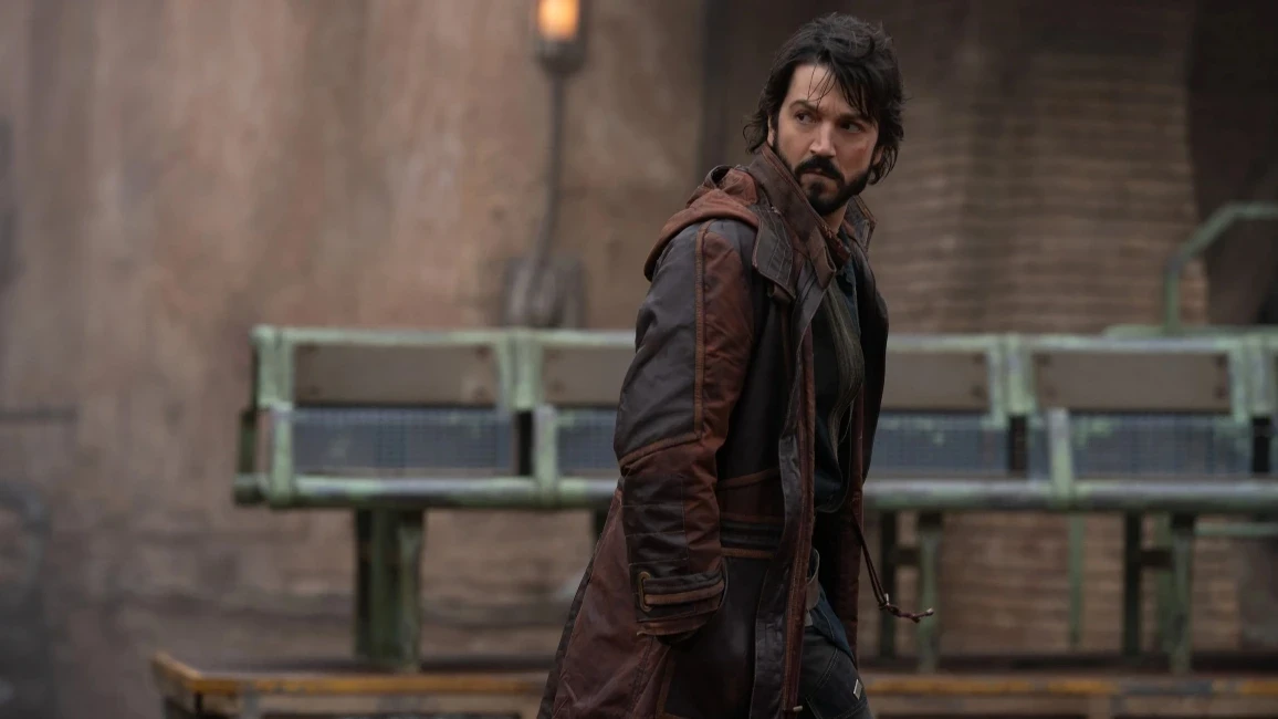 A man with dark hair and a beard stands in a rugged environment, wearing a long brown coat and dark clothing. They have a focused expression and are slightly turned to the side. A blurred industrial backdrop is visible.