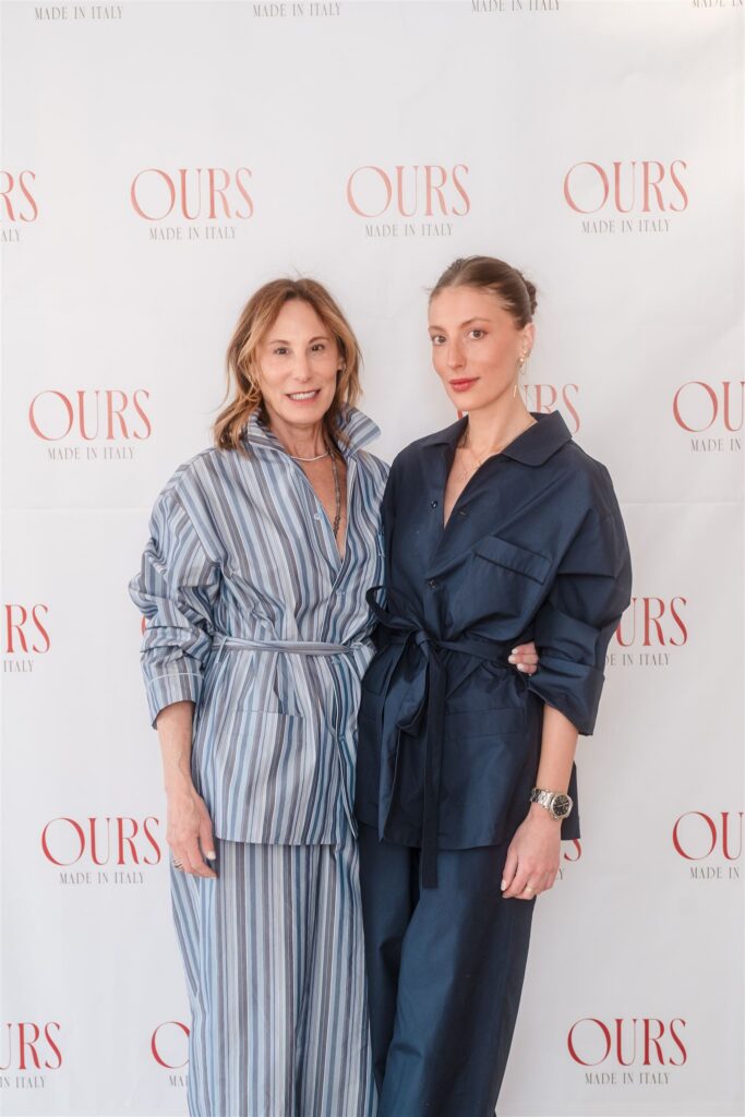 Two women posing in front of a branded "OURS Made in Italy" backdrop, showcasing Aspen luxury corporate event ideas with chic, high-end loungewear and striped coordinated sets.