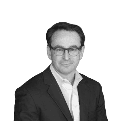 A person wearing glasses and a dark suit jacket over a light-colored shirt is seated against a teal background.