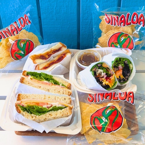 桌子上有各种三明治、卷饼,和装在品牌“锡那罗亚”袋子里的薯条。背景是蓝色的。