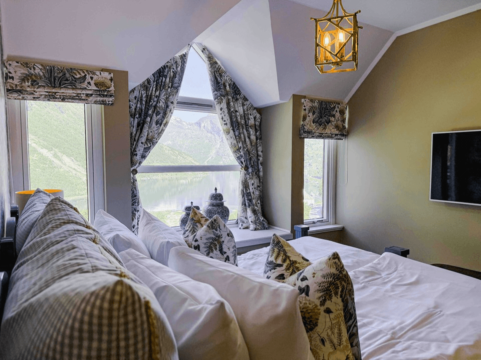Luxurious bedroom with a stunning view features a plush bed adorned with patterned pillows, elegant floral curtains framing large windows overlooking green mountains and a serene lake, and a stylish pendant light hanging from the ceiling.