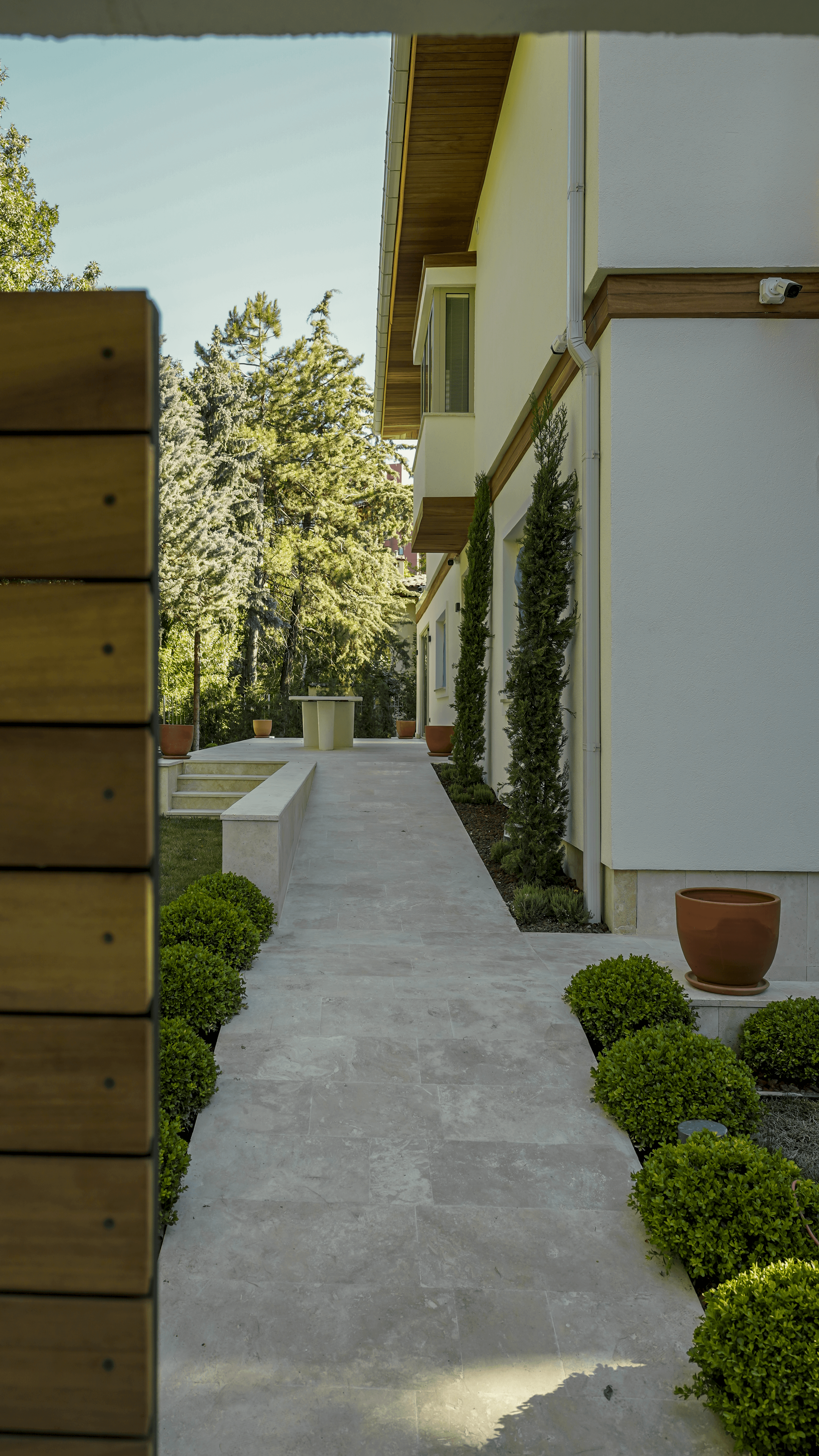 Serene garden walkway beside a modern villa with travertine flooring, vertical greenery, and wooden eaves detail under natural daylight.