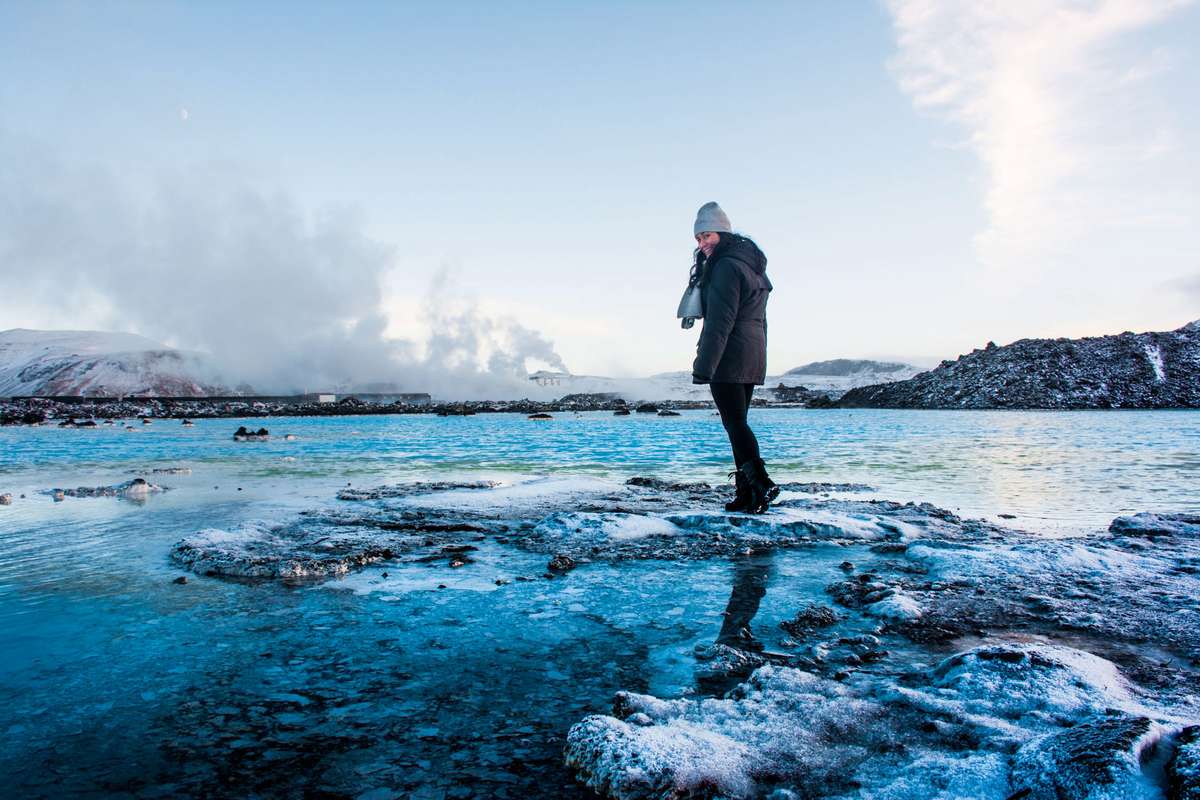 A person in dark winter clothing and a white beanie stands on a frozen lake with turquoise ice and snow.