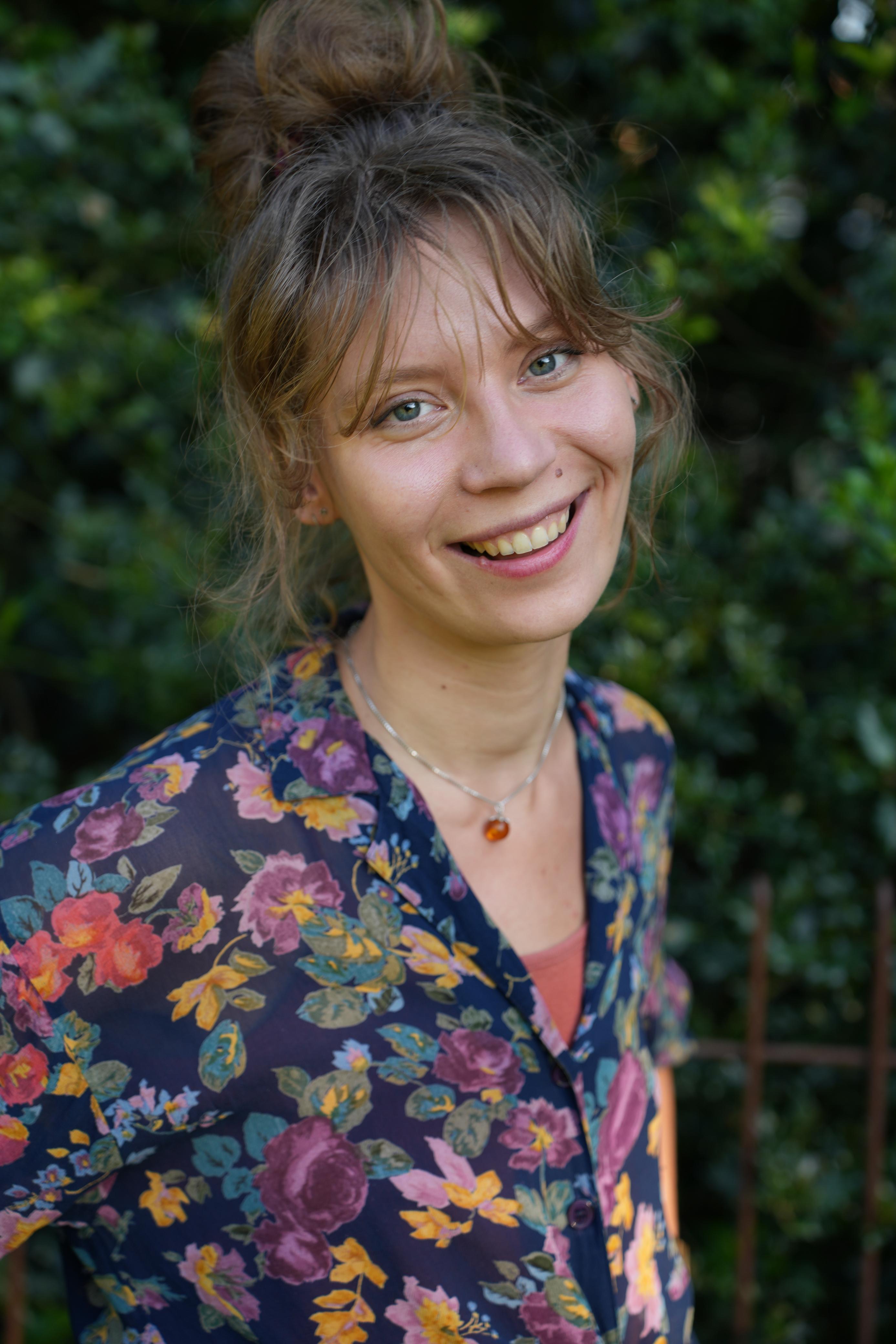 A smiling person with light brown hair in a bun is wearing a floral-patterned shirt with vibrant colors, standing in front of a lush, green foliage background.