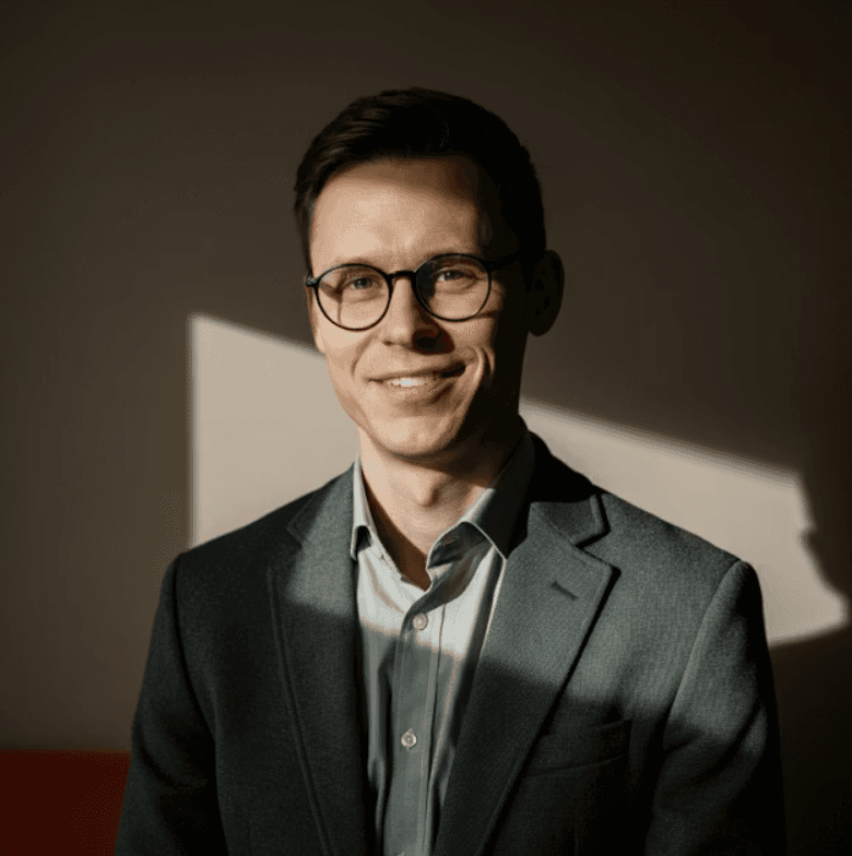 A professional portrait of a man with short hair, glasses, and a neatly trimmed beard, wearing a collared dress shirt and a blazer against a dark, textured background.