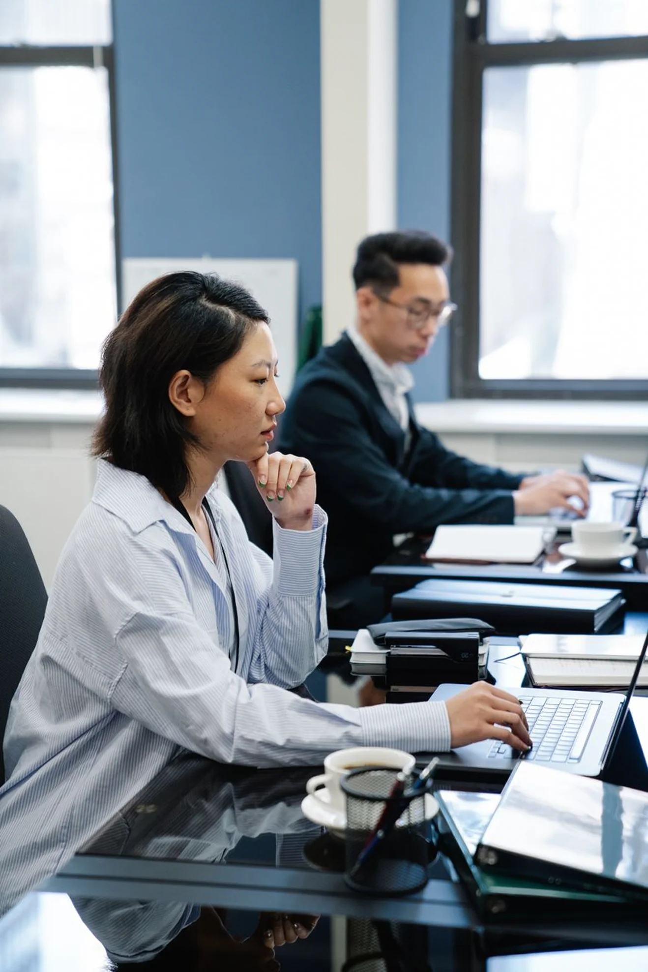 An Asian woman with a focused expression sitting at an office desk and working on her laptop. She is wearing a light blue striped button-down shirt, with her right hand typing and her left hand resting under her chin in deep thought. In the background, a male colleague wearing glasses and a dark suit jacket is also typing on his laptop at his desk. Coffee cups and office supplies are visible on both desks.