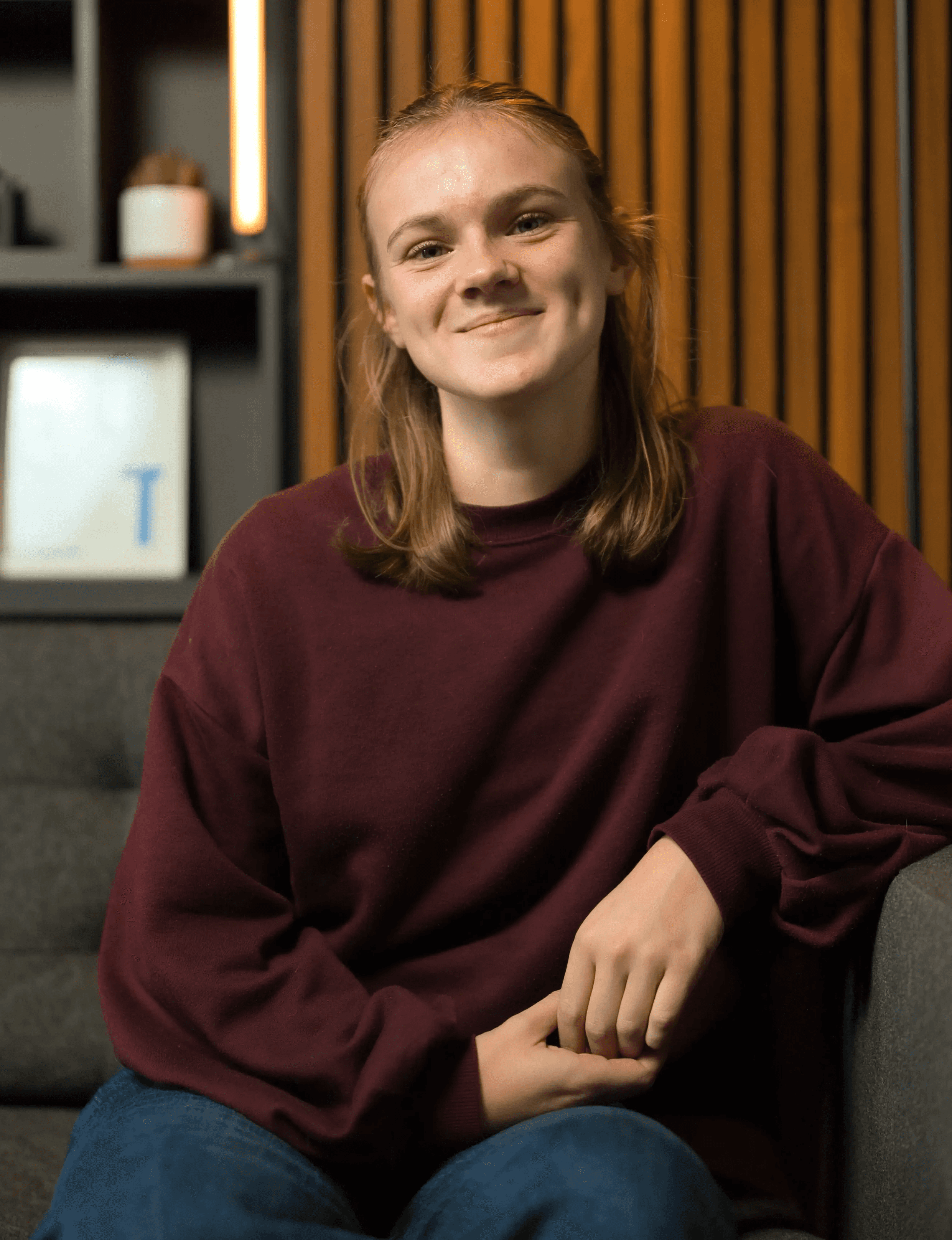 A person with shoulder-length hair, wearing a maroon sweater, is seated on a gray sofa with a warm, welcoming smile, set against a backdrop of wooden paneling and modern decor.