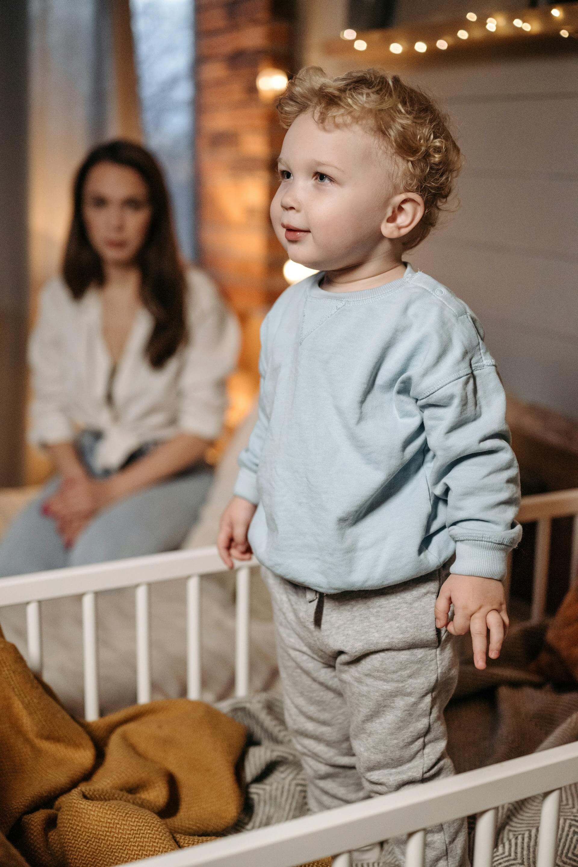 A young boy stands in a crib, smiling, while his mother watches lovingly in the background