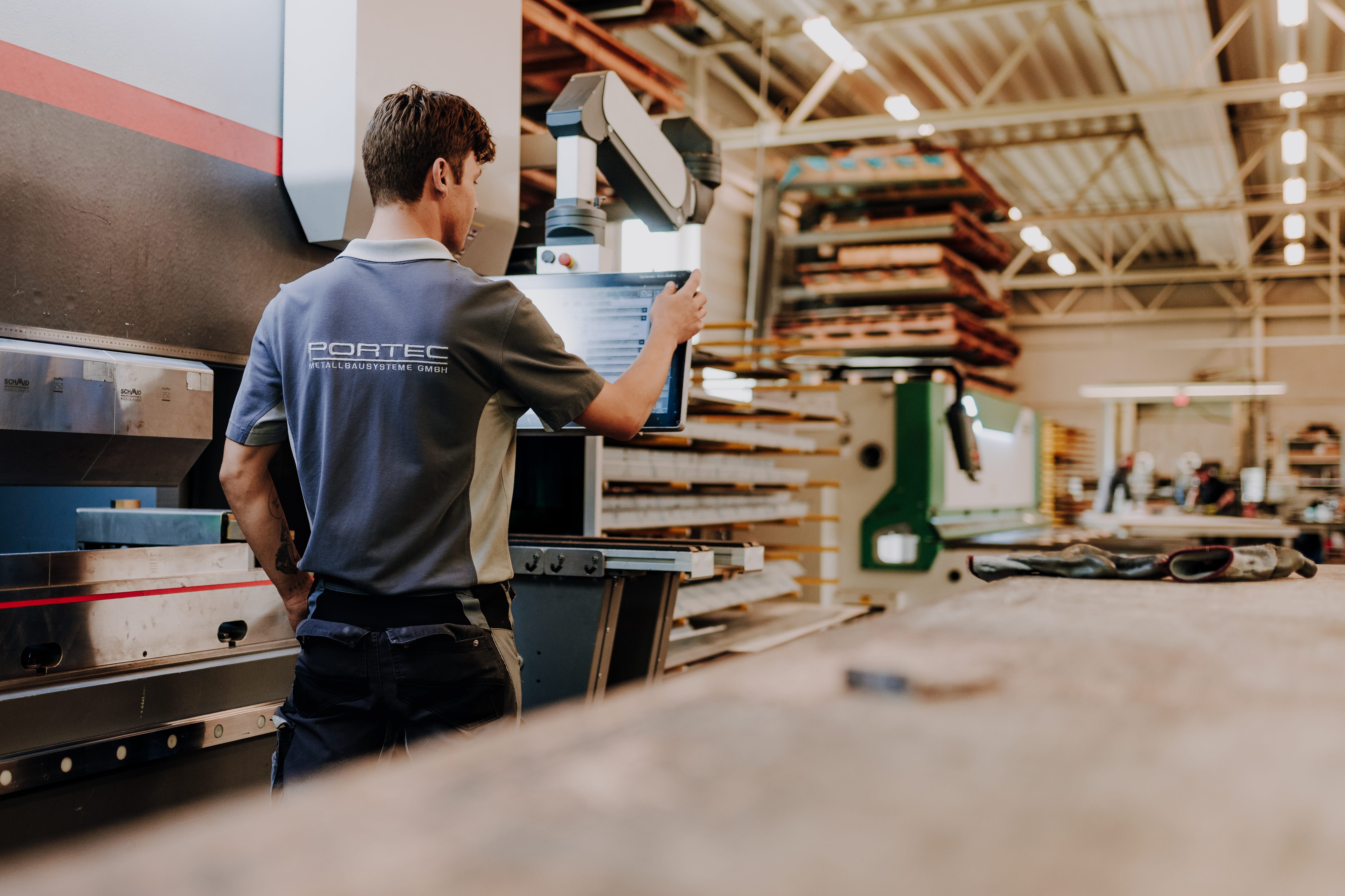 Man in a blue and gray shirt using a machine. A few machines and a stack of wood are visible.