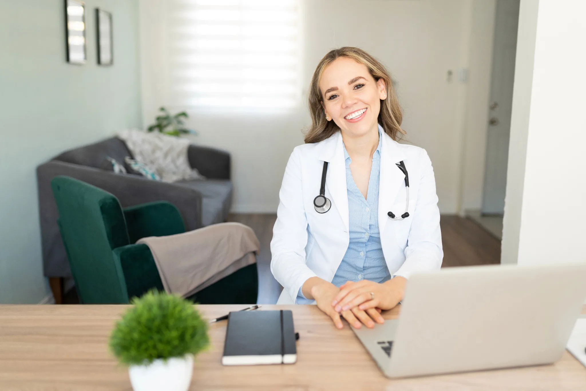 Smiling female physician in white coat with stethoscope around her neck sitting in front of her laptop
