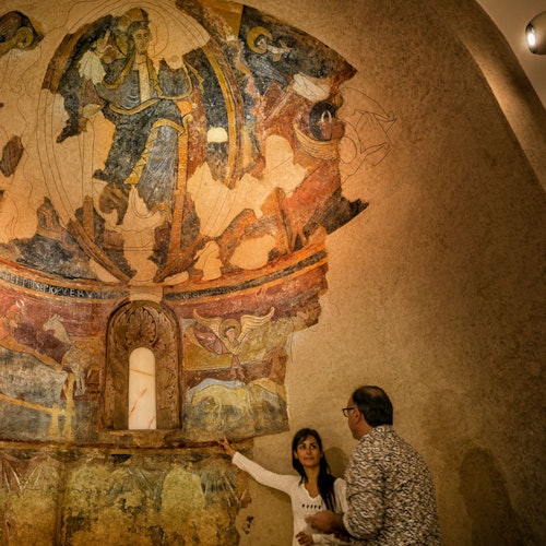 Two people observe a detailed mural of religious figures on the curved ceiling of a dimly lit room.