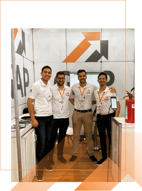 Quatro membros do time da GAP Sistemas, vestindo camisas polo brancas com o logo, sorrindo e posando no stand da empresa em um evento, com o logo da GAP ao fundo.