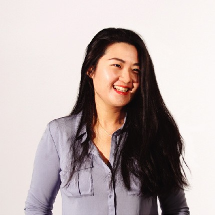 A woman with long, dark hair stands against a plain white background. She wears a light purple, button-down shirt with long sleeves and two chest pockets. Her head is turned slightly to the right, and she has a warm smile with her mouth open, revealing teeth. She is looking directly at the camera.