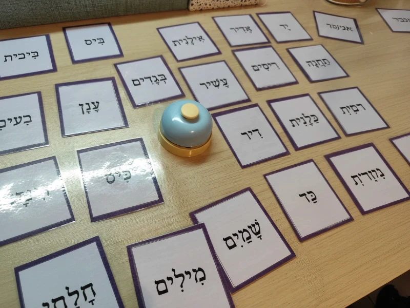 כרטיסיות עם מילים בעברית מנוקדת (כגון שמים, מילים, בגדים) פרוסות על שולחן לצד פעמון שולחן כחול למשחק לימודי בקליניקה של סמדר עציץ נעים ולומדים