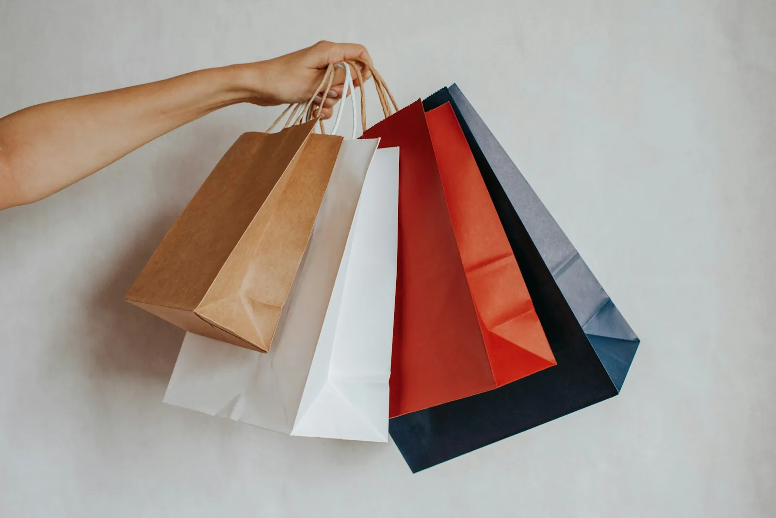 Outstretched arm holding a bundle of kraft, white, red and navy paper shopping bags against a pale grey wall