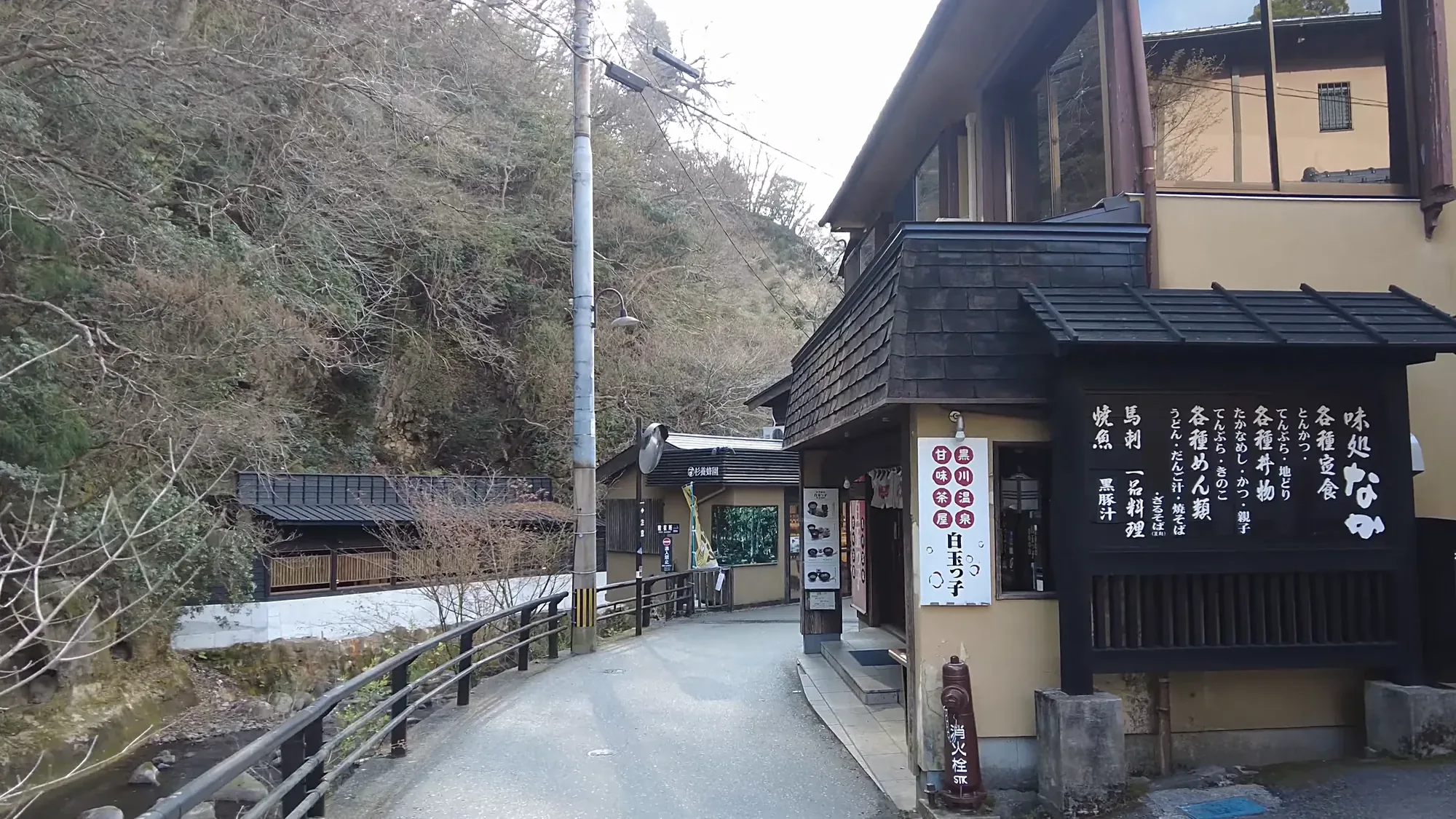 A lane of dark timber shopfronts with hanging signs at Kurokawa Onsen, a hill behind