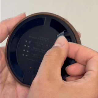 Hands peeling off the protective film from the adhesive side of a round, black ZenStand Adhesive Base, preparing it for placement.