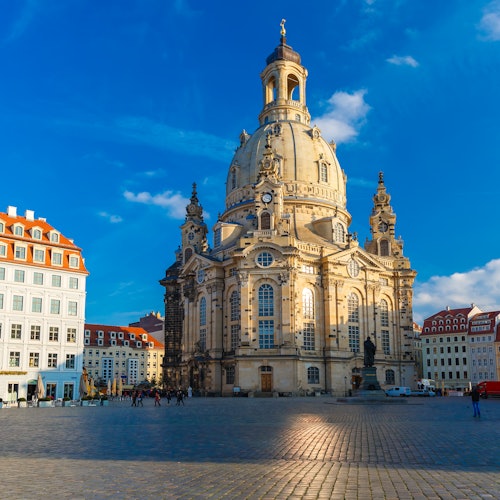 Aurinkoisella aukiolla seisoo suuri barokkityylinen kirkko, jonka keskellä on kupoli. Kirkkoa ympäröivät pienemmät rakennukset ja muutama ihminen.