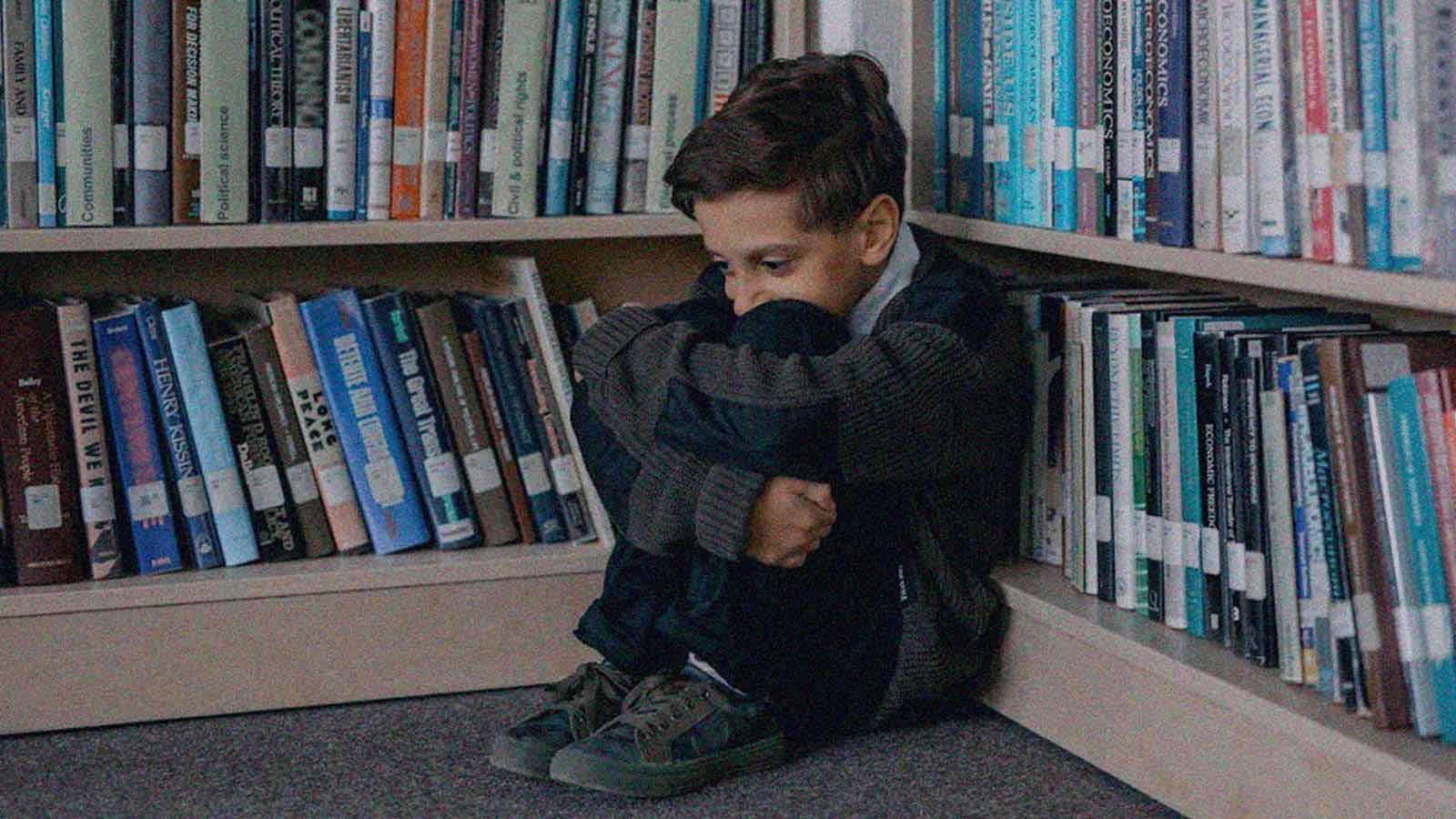 Foto de un niño sentado en el suelo de una biblioteca mientras se sostiene las piernas y oculta su rostro en la columna de opinión Si un niño dice que sí, es no de IdeaPaís.