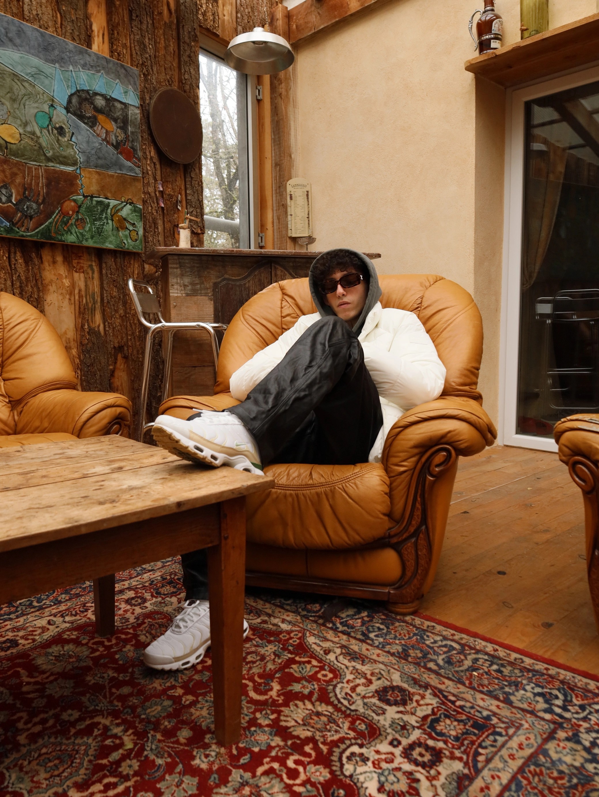 Person in sunglasses and hoodie sits on a tan leather chair in a rustic room with wooden walls and a vintage rug.