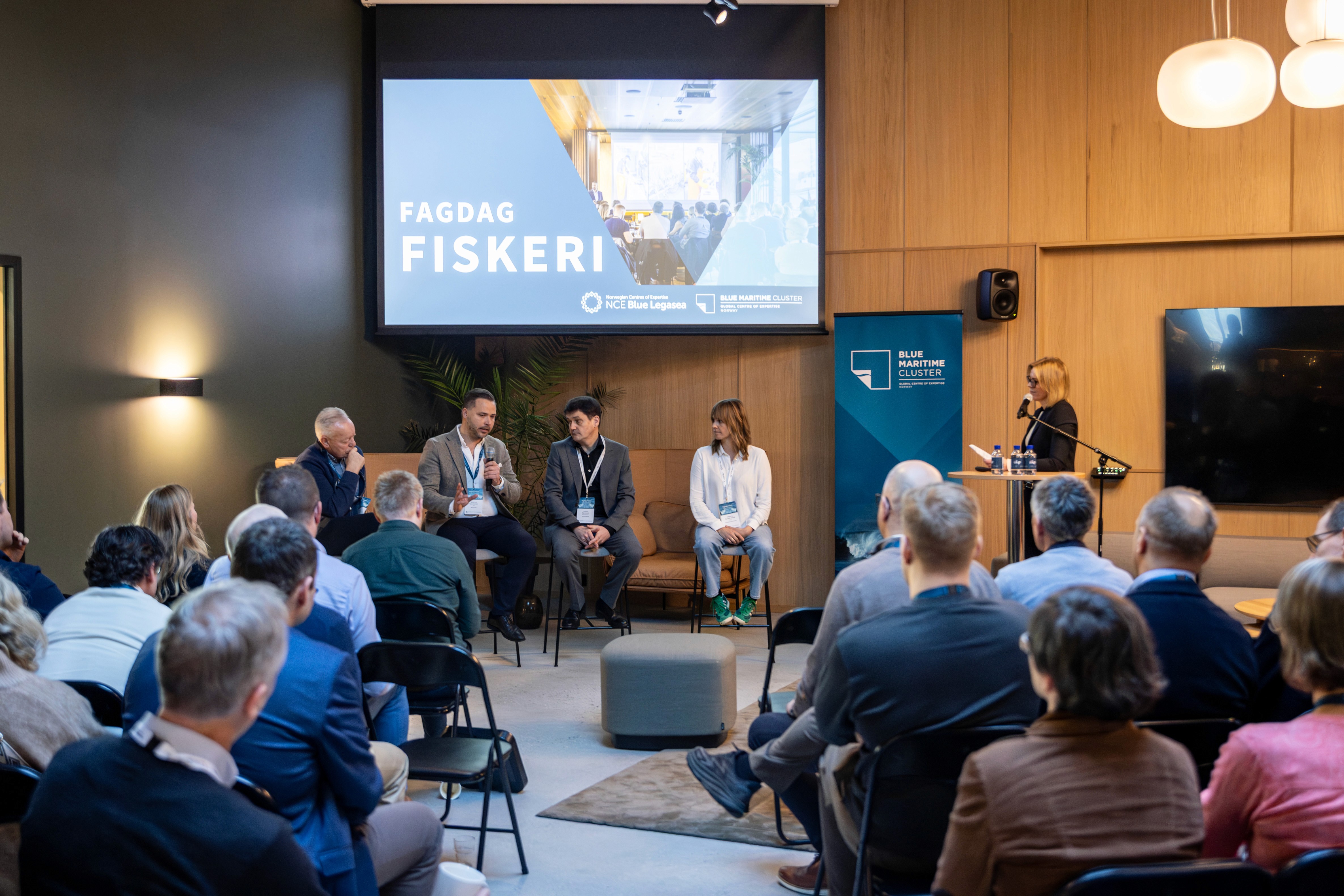 A presentation in progress with a speaker at the front, audience facing a large screen.