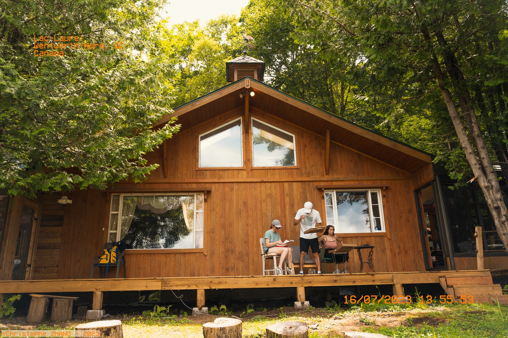 Terrasse d’un chalet au bord d’un lac, trois amis discutent sous un ciel d’été.
