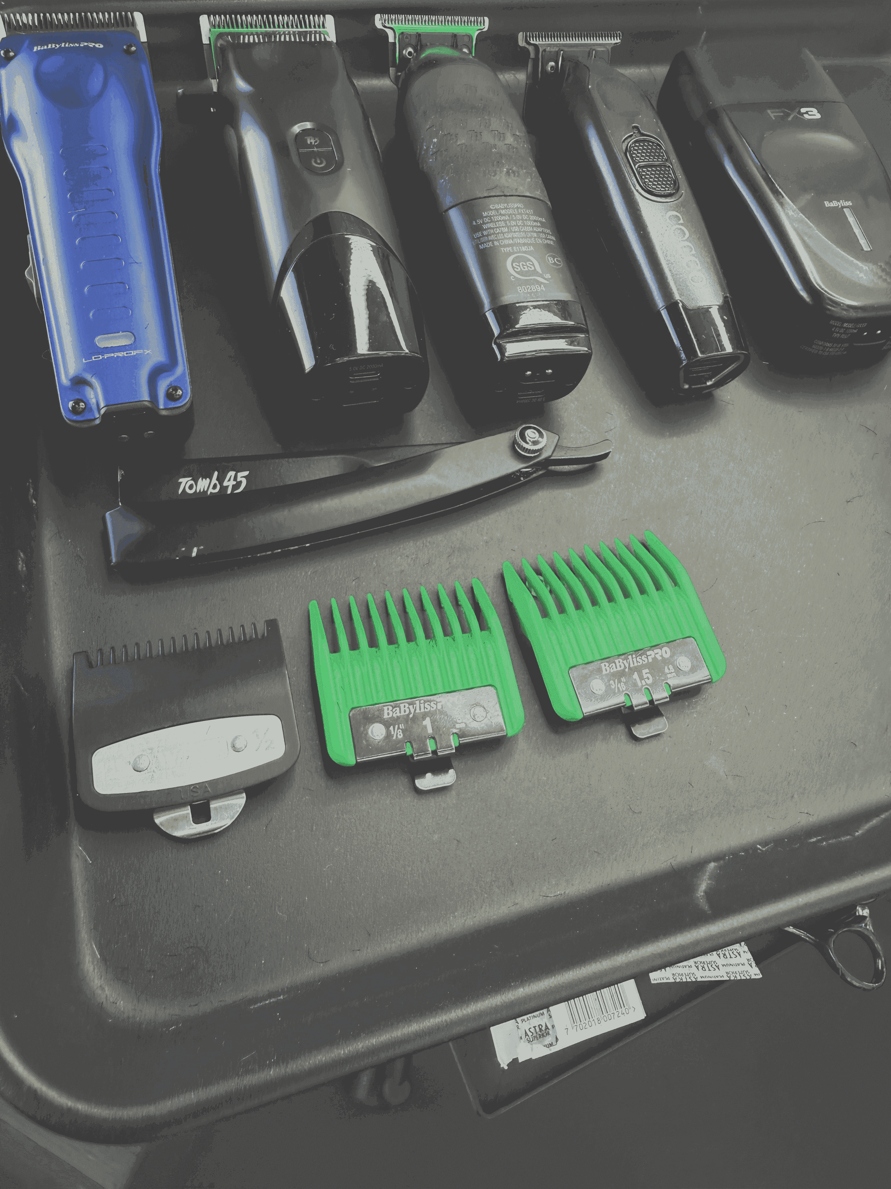 Close-up of barber tools on a workstation, including clippers, trimmers, a straight razor, green clipper guards, and other grooming accessories.